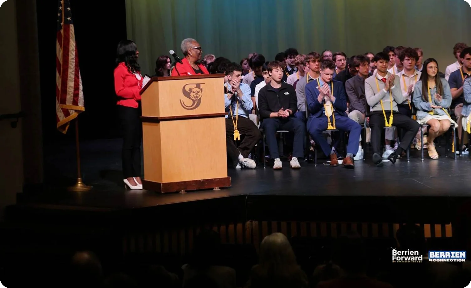 A speaker at a lectern on a school stage with students seated behind her, applauding